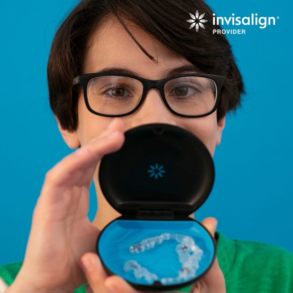 A person with short dark hair and glasses holds an open Invisalign case with clear aligners inside, against a bright blue background. The Invisalign logo appears in the top right corner.