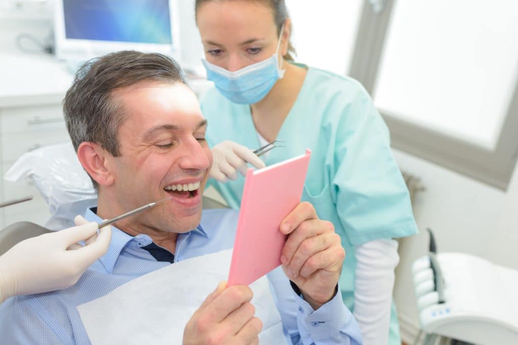 A man sits in a dental chair smiling and looking at his reflection in a pink hand mirror, while a dental professional in scrubs and a mask examines his mouth with dental tools.