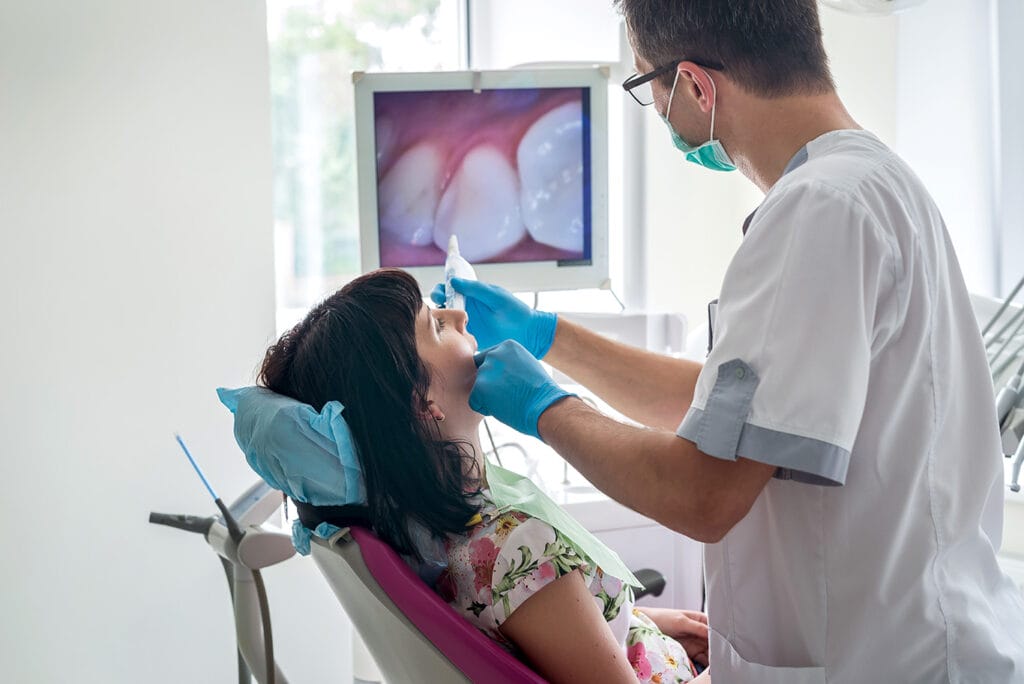 A dentist wearing gloves and a mask examines a woman’s teeth in a dental clinic, while an image of her teeth is displayed on a monitor in the background—showcasing the importance of dental diagnostic services for oral health.