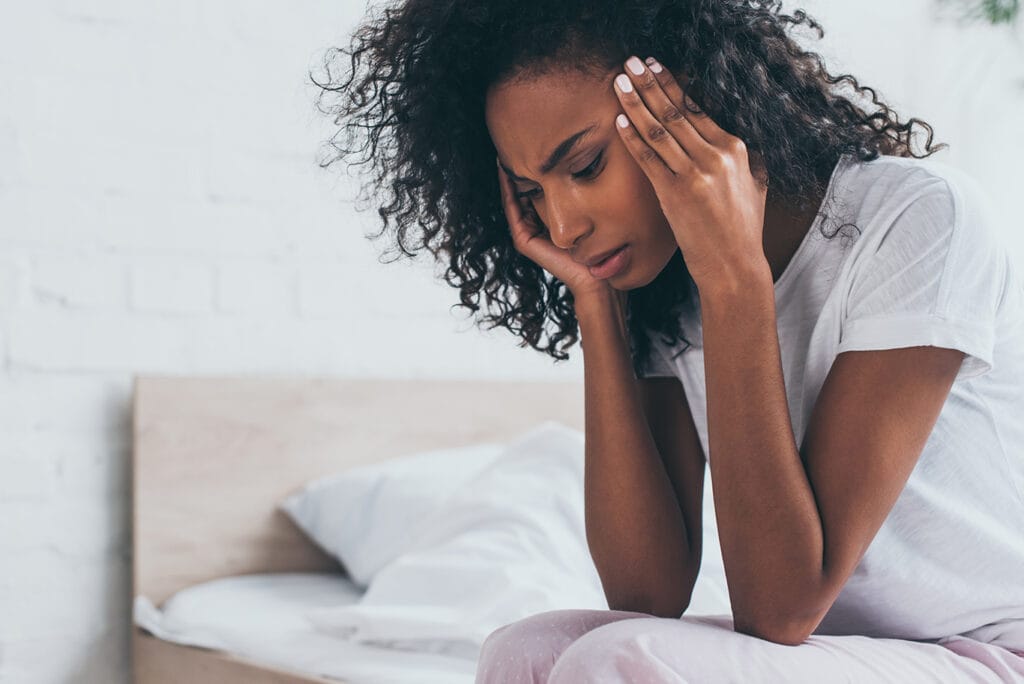 A woman sits on the edge of a bed, holding her head in her hands and appearing distressed or overwhelmed, possibly contemplating upcoming dental medical treatments. She is wearing a white t-shirt and pink pants, with an unmade bed in the background.
