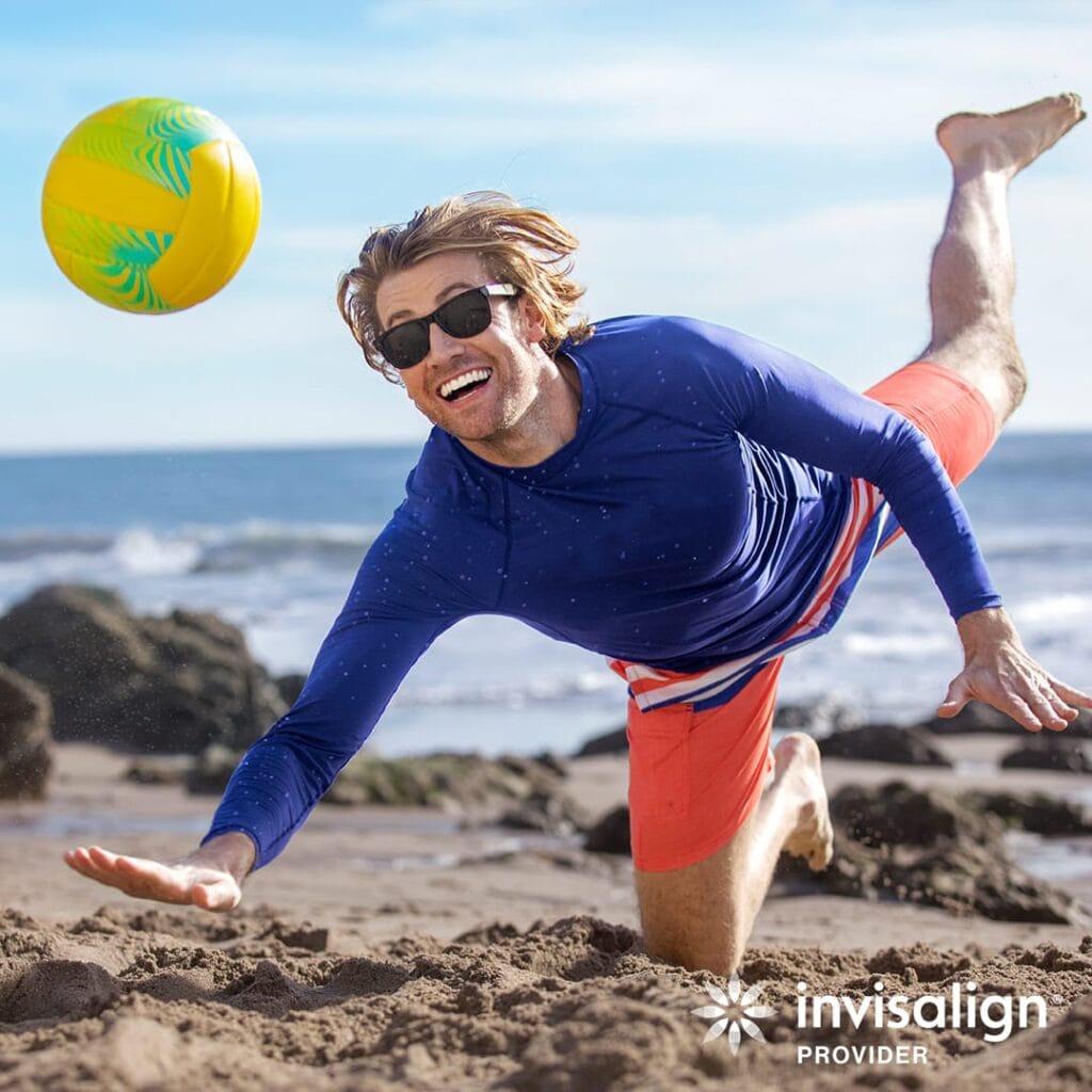 A man in sunglasses and swimwear dives on a sandy beach to hit a colorful volleyball, smiling energetically, with the ocean and rocks in the background. The "Invisalign Provider" logo appears in the corner.
