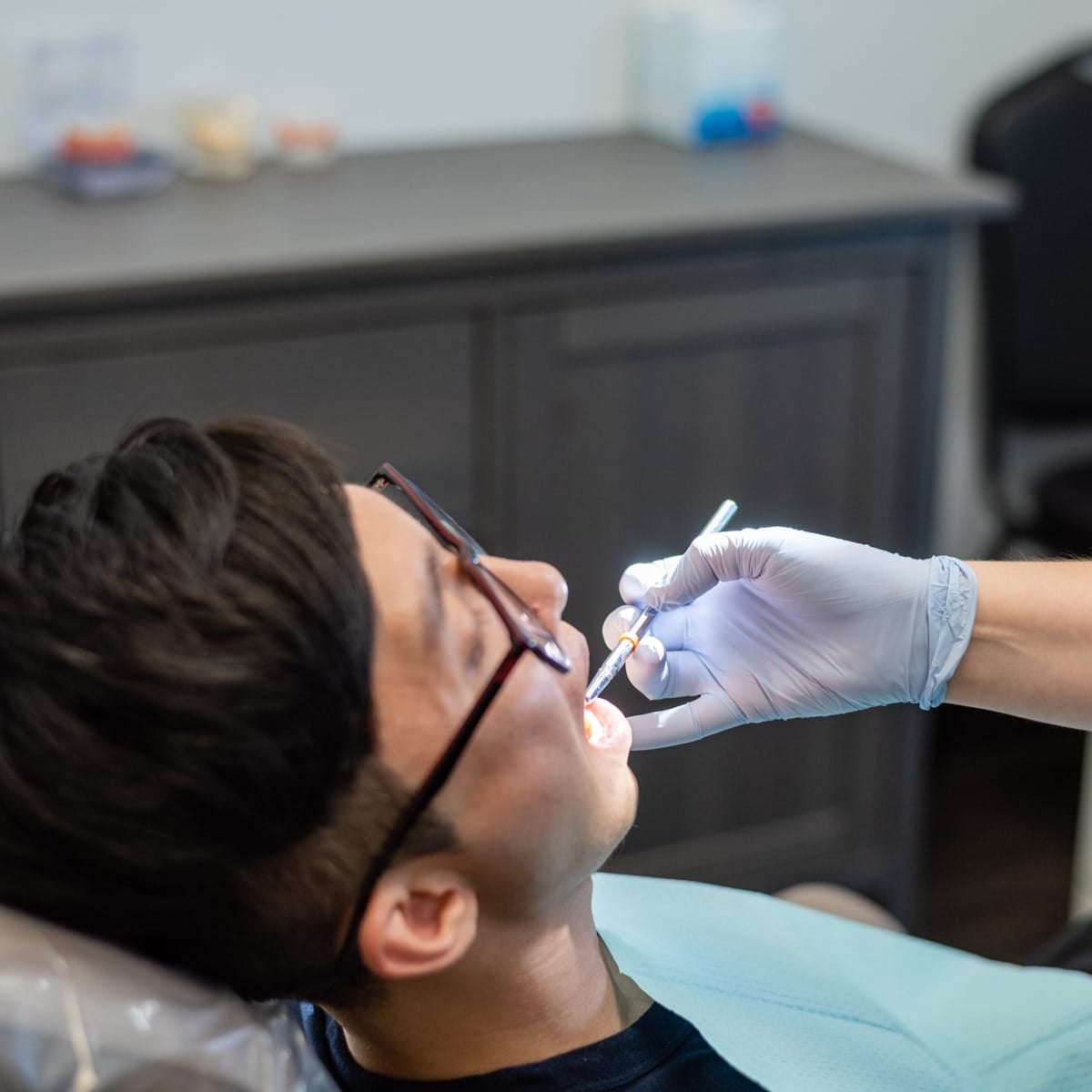 Patient wearing glasses reclines in a dental chair while a gloved dental professional uses an instrument inside the patient’s mouth
