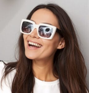 A smiling woman with long brown hair wears white, oversized sunglasses printed with bird eyes on the lenses. Dressed in a white top, she appears happy and playful, much like someone who just left glowing Citrus Heights dental reviews.