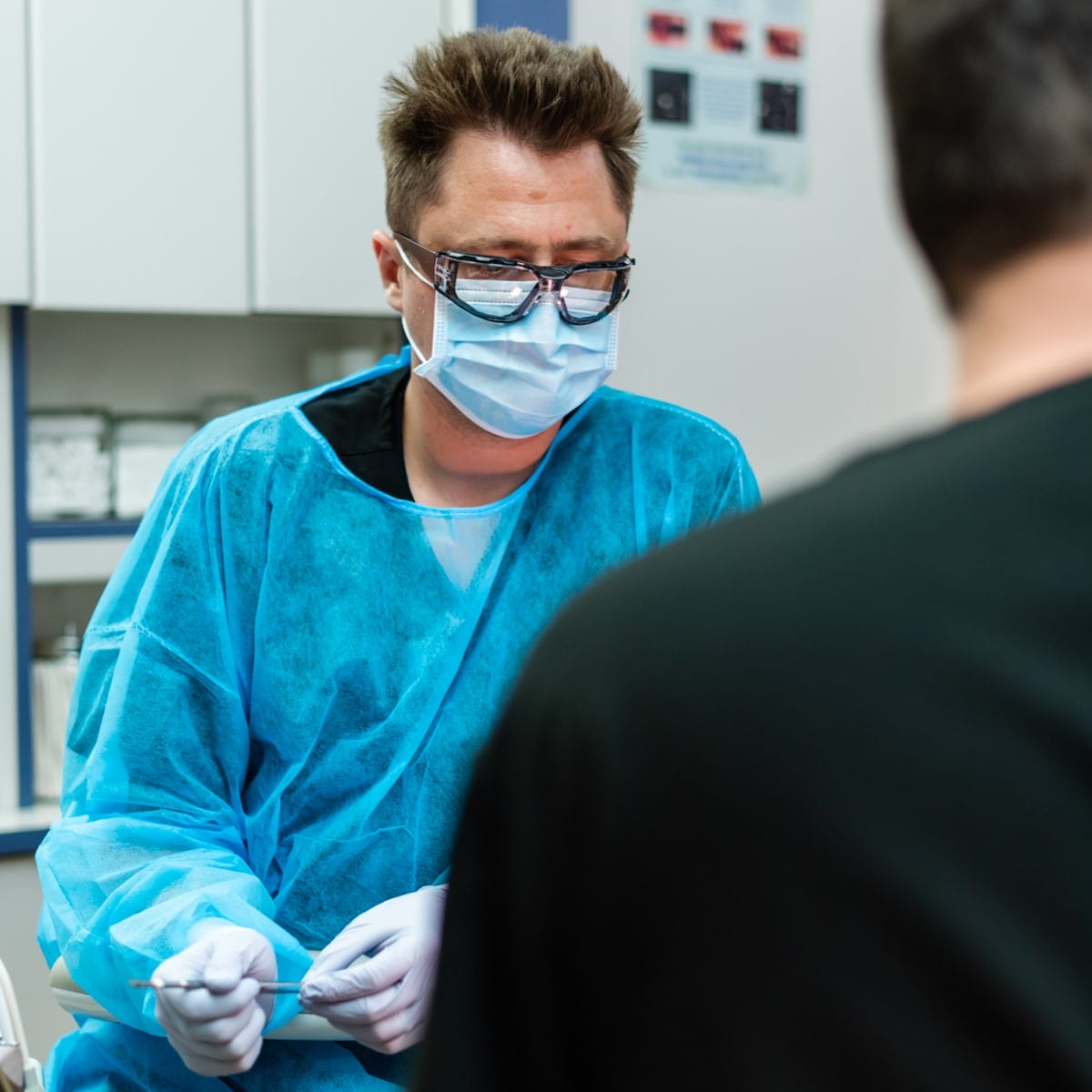 A healthcare worker in a blue gown, mask, and gloves sits with medical tools, speaking to a seated patient in a clinic setting. Shelves and medical posters are visible in the background.
