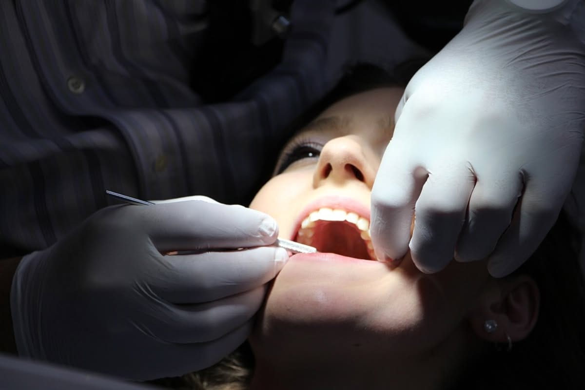 A person wearing white gloves examines a patient's mouth with dental tools while the patient lies back with their mouth open, during a dental check-up.