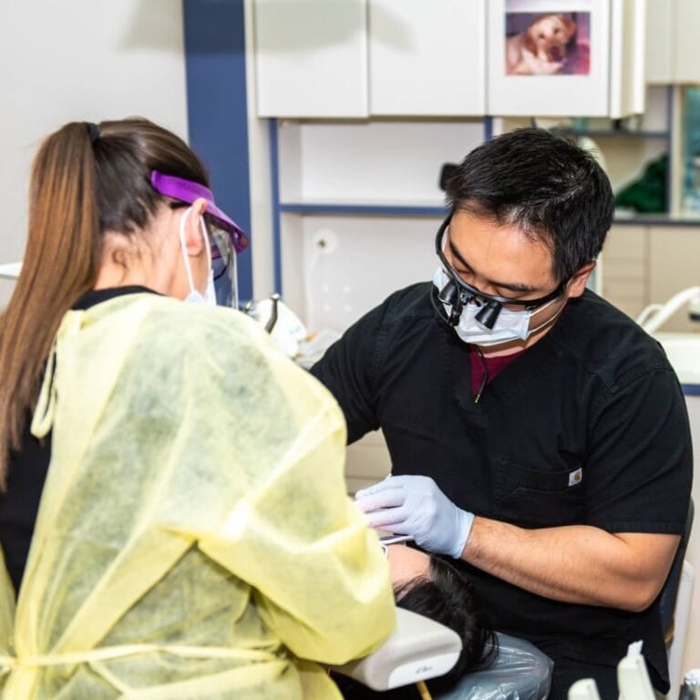 A dentist wearing protective glasses and a mask works on a patient’s teeth, assisted by a dental hygienist in a yellow gown and face shield, in a brightly lit dental clinic.
