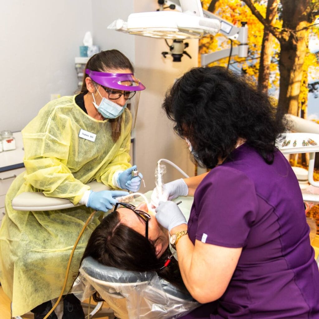 Two dental professionals, one in yellow PPE and face shield and the other in purple scrubs, treat a patient lying in a dental chair in a brightly lit office with a nature-themed wall mural.