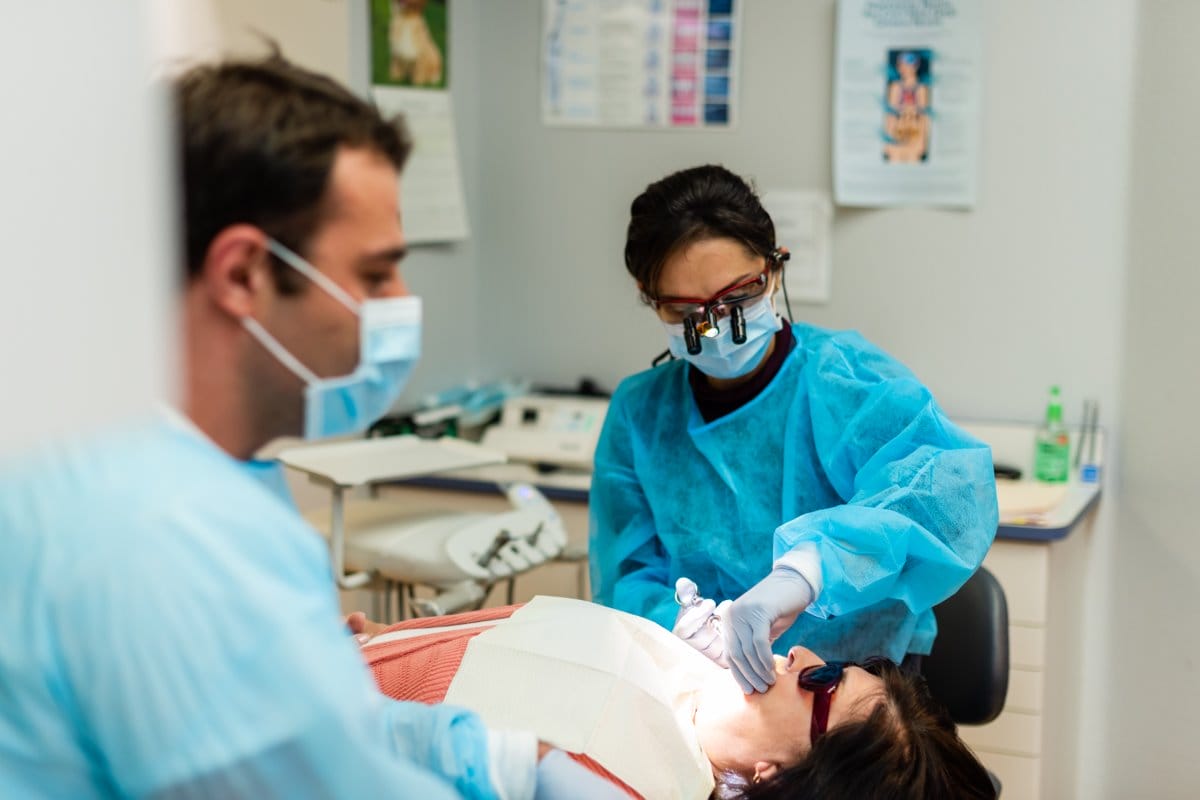 A dentist and assistant, both wearing blue gowns, masks, and gloves, perform a dental procedure on a patient lying back in a dental chair under bright lights in a clinic room.
