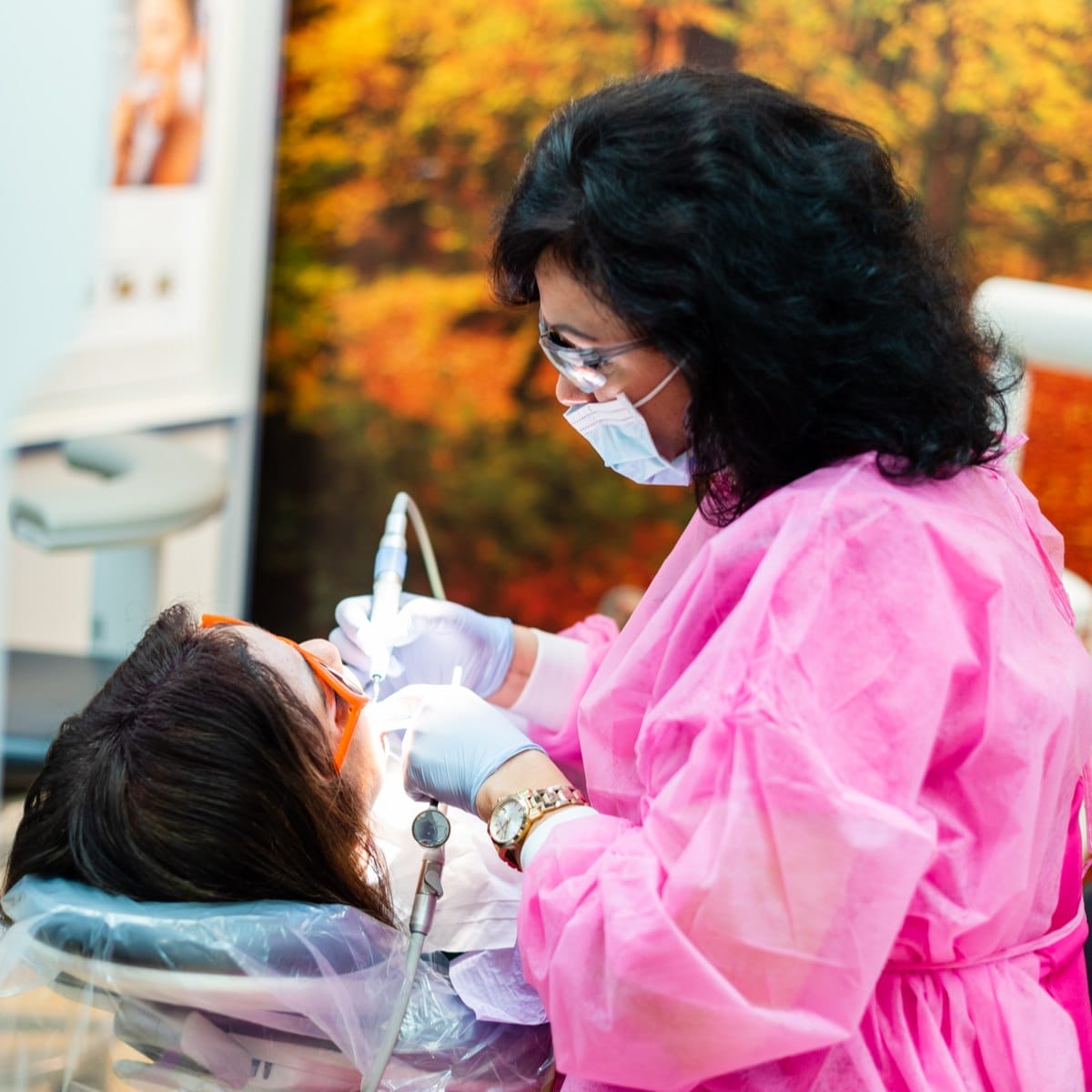 A dentist wearing a pink gown, gloves, face mask, and goggles works on a patient lying back in a dental chair. The patient wears protective glasses, and an autumn forest image is in the background.