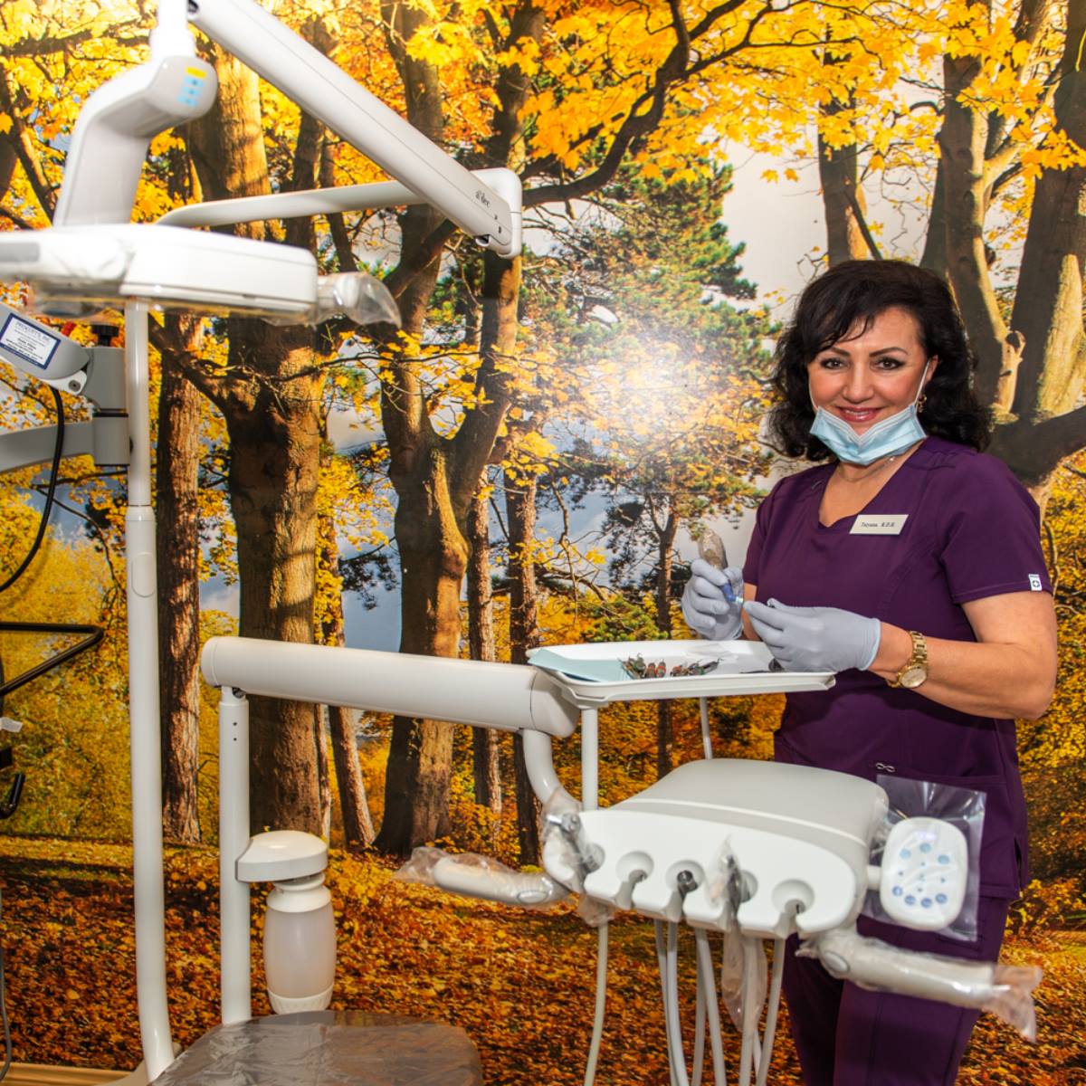 A female dentist in purple scrubs and gloves stands in a dental office, smiling at the camera. Behind her is a mural of a bright autumn forest with yellow leaves. Dental equipment is visible in the foreground.