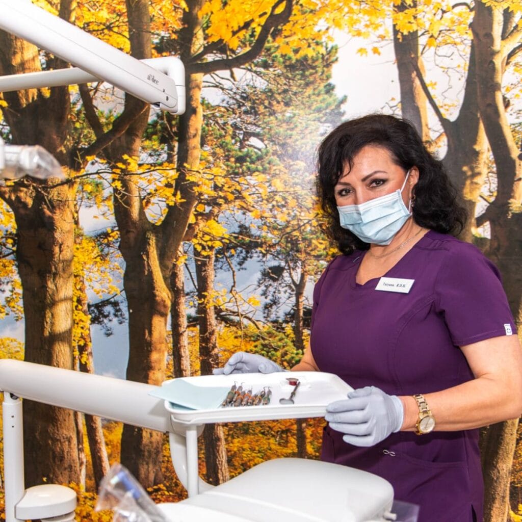 A female dental professional wearing a purple uniform, gloves, and a face mask stands in a dental office, holding a tray of dental tools. Behind her is a wall mural of trees with yellow autumn leaves.