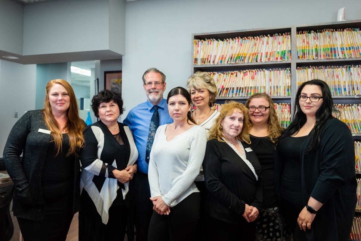 A group of eight adults, dressed in business attire, stand together smiling in an office with shelves of color-coded files in the background.