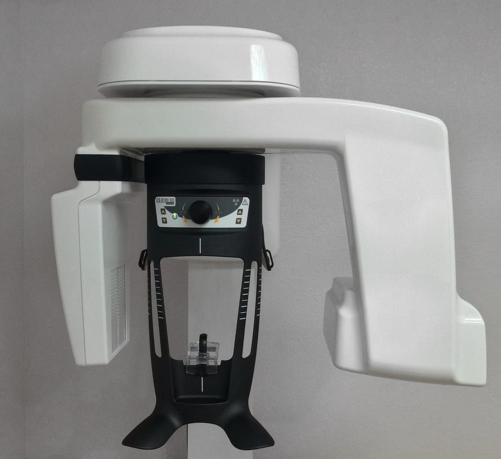 A panoramic dental X-ray machine with a white and black exterior, control panel, and chin rest, mounted against a light grey wall.