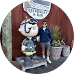 A person stands next to a large statue of a cartoon character dressed as a pilot, outside a wooden building with potted plants and a sign that reads "LIFT-OFF AIR," pausing to read several cheerful reviews posted near the entrance.