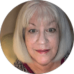 A woman with straight, shoulder-length gray hair and bangs smiles gently at the camera. She is indoors, wearing a patterned top, and appears ready to share her testimonial with patients seeking honest reviews.