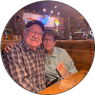 An older man and woman, likely patients, sit closely together at a wooden table in a cozy restaurant, smiling at the camera. Neon lights and rustic decor create a warm atmosphere perfect for sharing good reviews and memories.