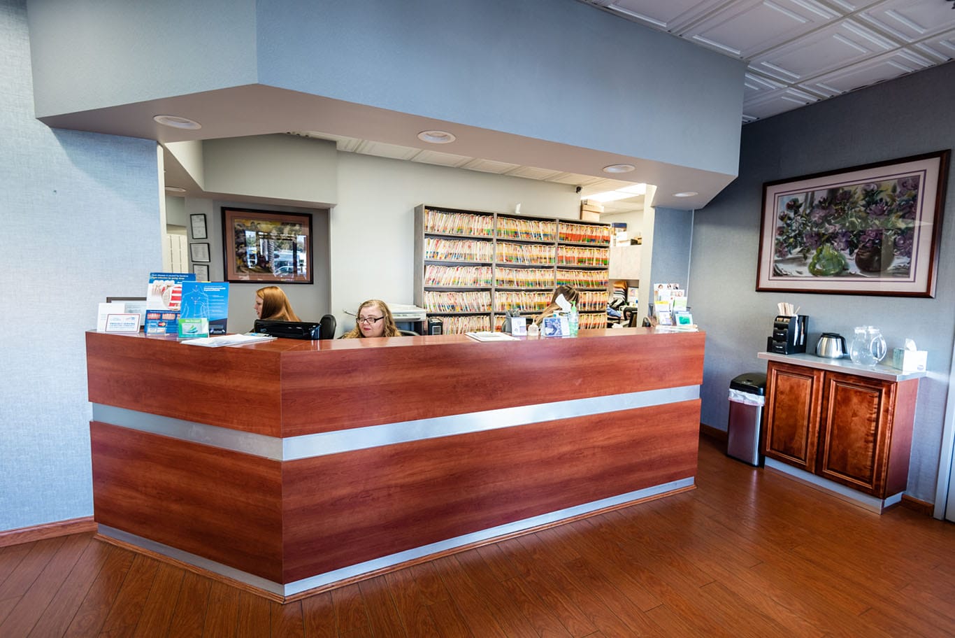 A modern medical office reception area with two women working at a wooden front desk, shelves of organized files behind them, and a coffee station with cups to the right.