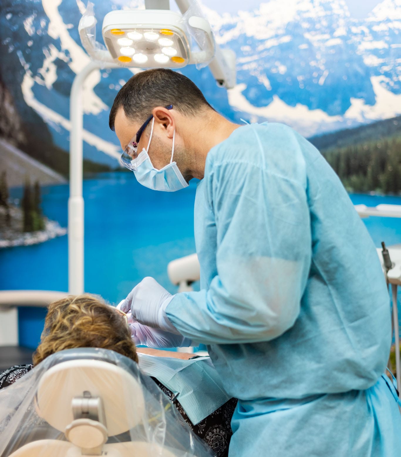 A dentist in a mask and blue gown examines a patient reclining in a dental chair. Dental equipment is visible, with a scenic mural of mountains and a lake on the wall behind them.