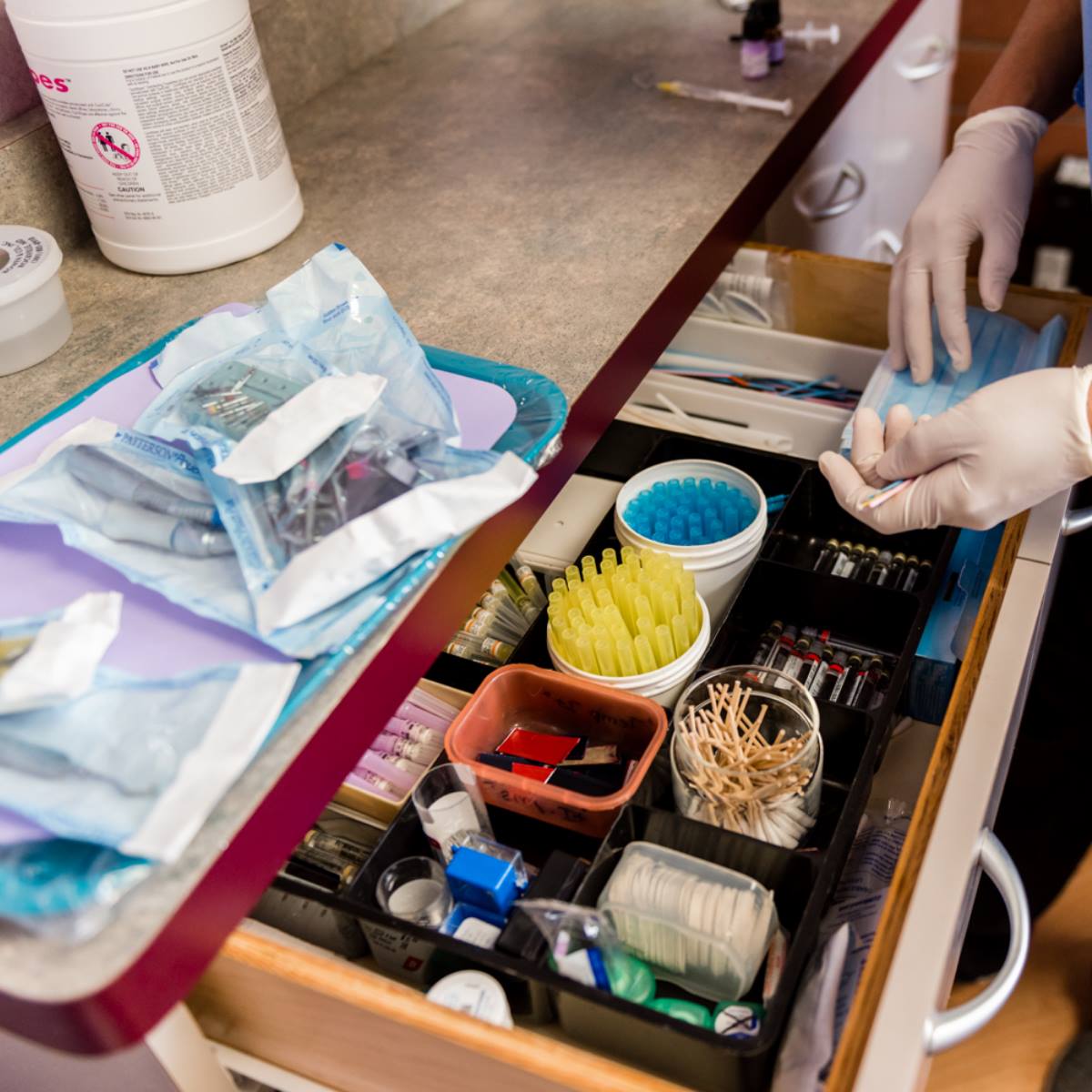 A person wearing gloves prepares dental tools next to an open drawer filled with various dental supplies, including syringes, swabs, and containers, in a clinical setting.