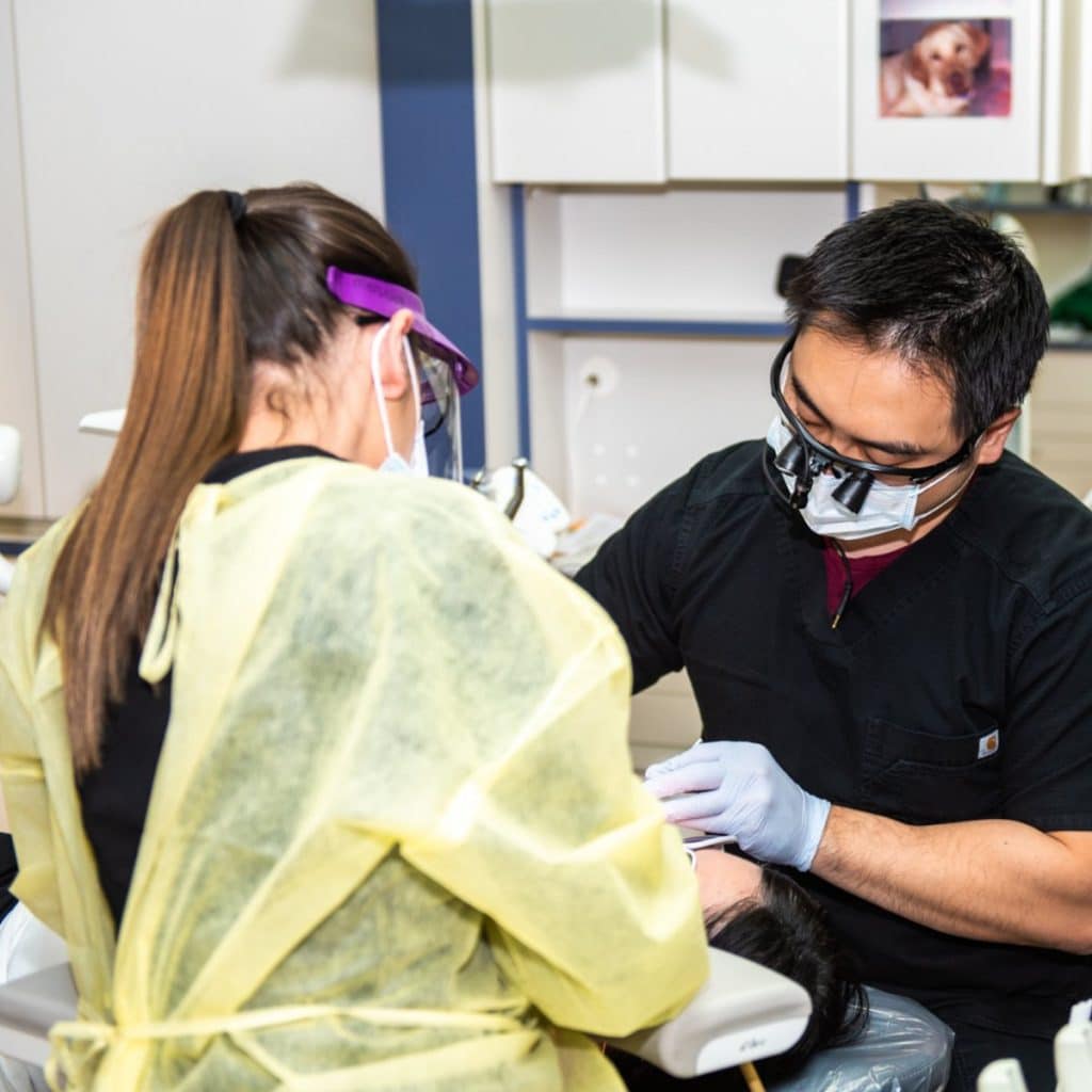 A dentist wearing a mask and gloves examines a patient's mouth while an assistant in a yellow gown and face shield assists in a brightly lit Sacramento dental clinic, reflecting our core values of care and professionalism.