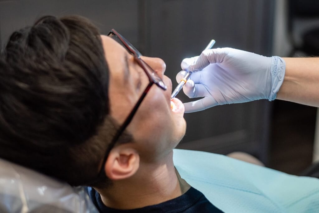 A person wearing glasses lies in a dental chair while a dentist with gloved hands examines their mouth using dental tools.