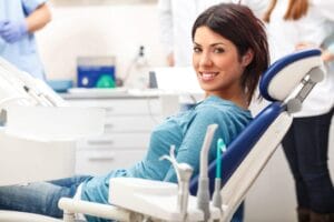 A young woman with dark hair, wearing a blue shirt, smiles while sitting in a reclining dental chair in a bright dental office, with dental tools and staff visible in the background.