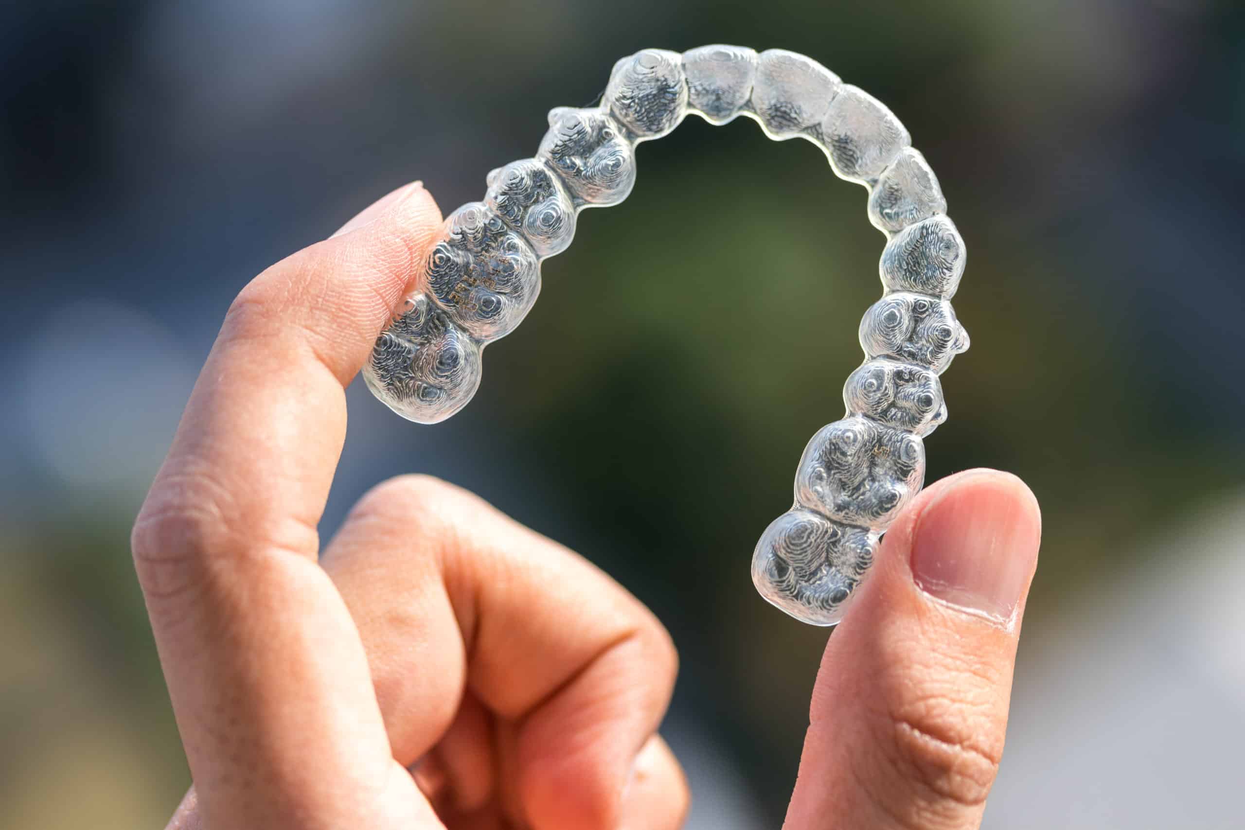 A hand holding a clear dental aligner against a blurred background. The aligner is transparent and shaped to fit over teeth, commonly used for orthodontic treatment.